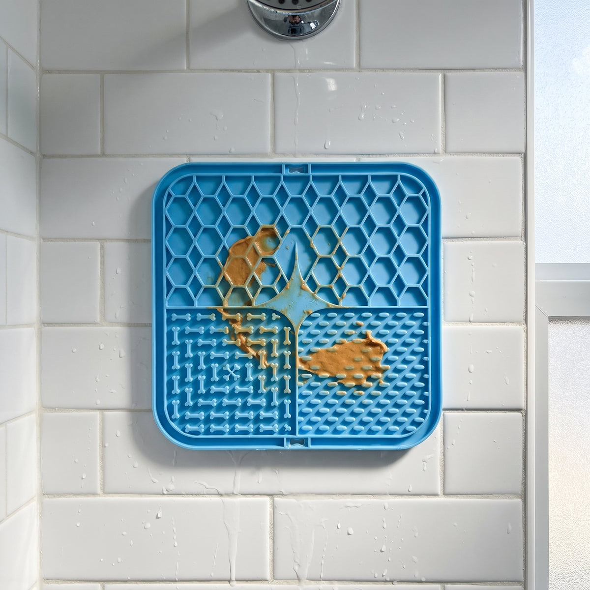 Blue lick mat stuck flat to a white shower tile wall with a smear of peanut butter