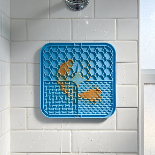 Blue lick mat stuck flat to a white shower tile wall with a smear of peanut butter