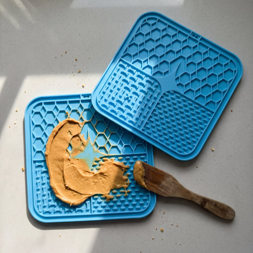 Two blue suction lick mats on a kitchen counter, one with peanut butter spread on it and a wooden spatula
